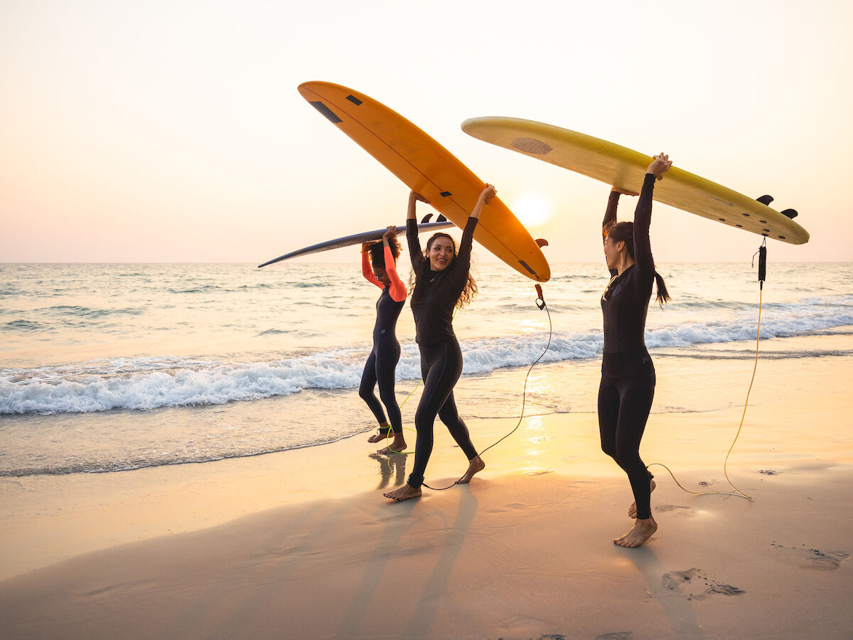 Four people wearing wetsuits carry surfboards on a beach at sunset, heading toward the ocean as waves gently hit the shore.