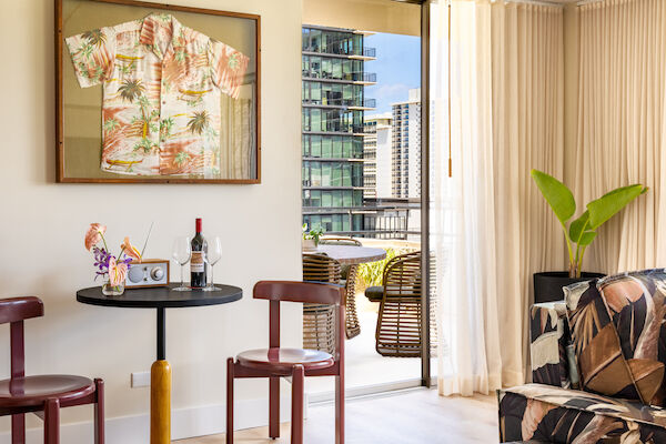 A cozy room with a small table and two chairs, tropical decor, a framed shirt, wine, and a parrot statue, leading to a balcony with city views.