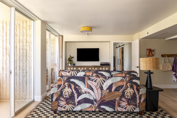 A living room with a patterned sofa, wall-mounted TV, sliding glass doors, and a checkered rug. The room has a cozy and modern decor style.