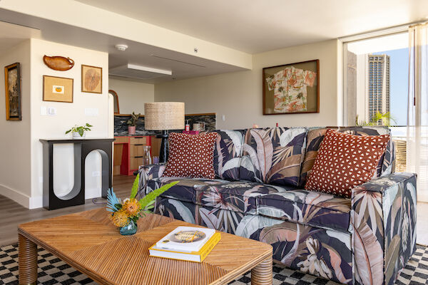 A cozy living space with a patterned sofa, two red polka dot pillows, a wooden coffee table, a decorative lamp, and a large window letting in sunlight.