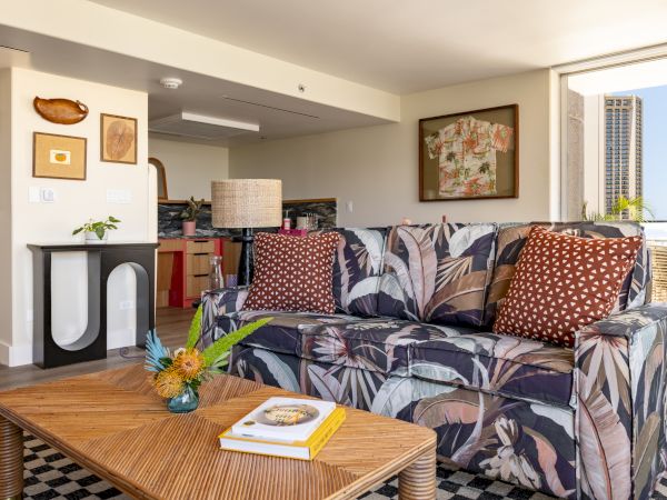 A cozy living space with a patterned sofa, two red polka dot pillows, a wooden coffee table, a decorative lamp, and a large window letting in sunlight.