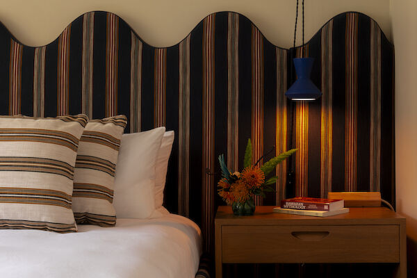 A cozy bedroom setup with a bed featuring striped pillows, a wooden nightstand with a flower vase, books, and a lamp in a blue shade hanging above.