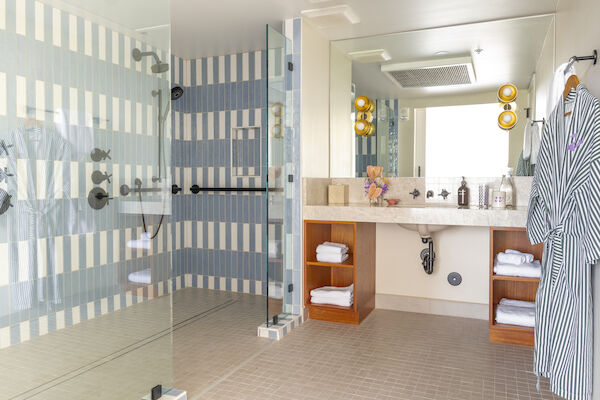 A modern bathroom features a large glass shower with striped tile, a double sink vanity with mirrors, towels, and a bathrobe hanging on the wall.