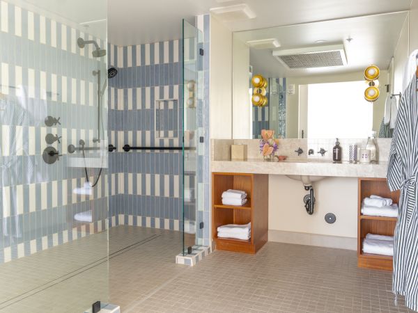 A modern bathroom features a large glass shower with striped tile, a double sink vanity with mirrors, towels, and a bathrobe hanging on the wall.