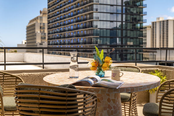 A rooftop patio with a round table featuring a magazine, a vase of flowers, a carafe of water, and a mug. Buildings are in the background.