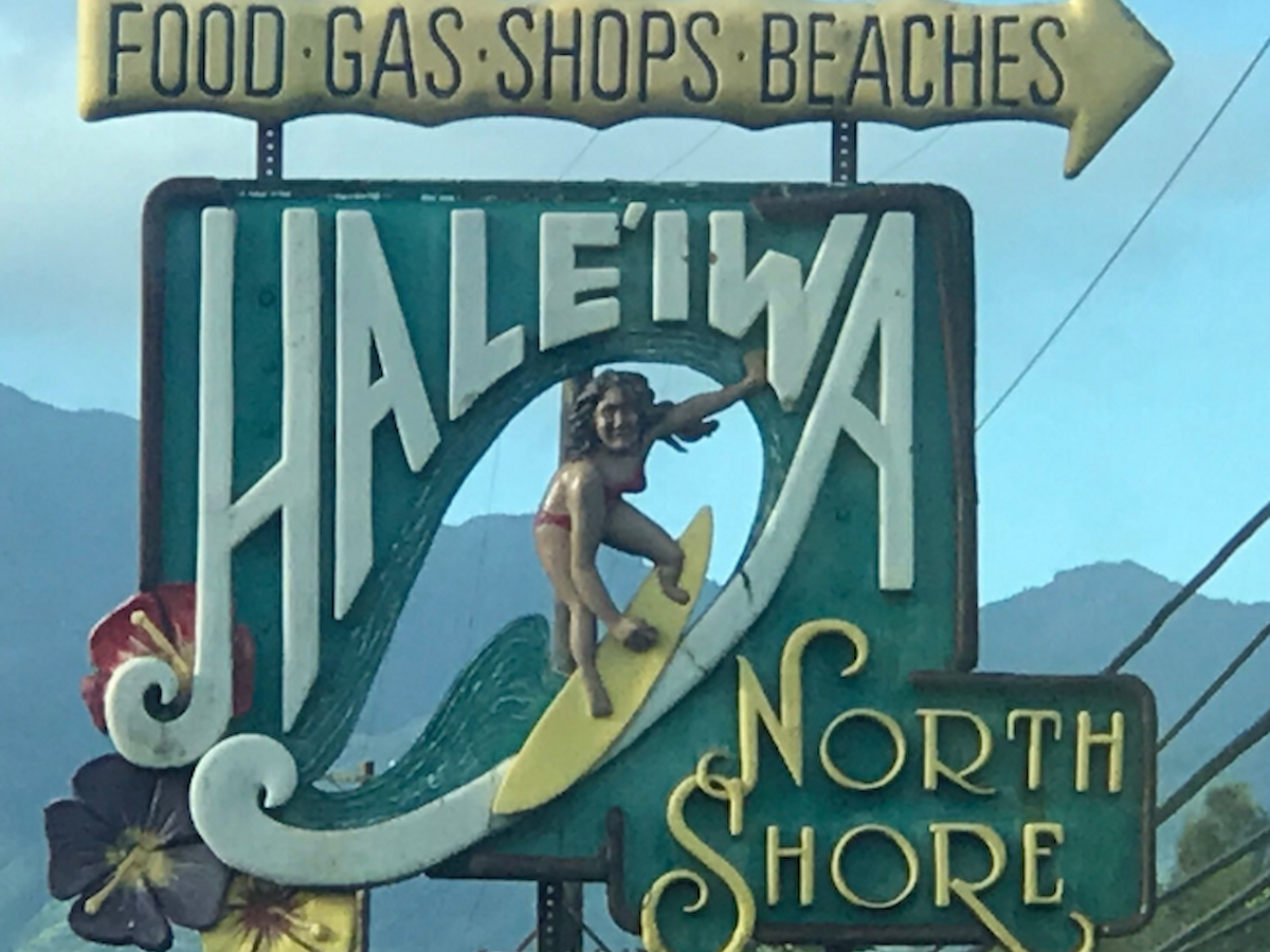 This is a sign for Haleiwa North Shore with a surfer depicted. It also points to food, gas, shops, and beaches, ending the sentence.