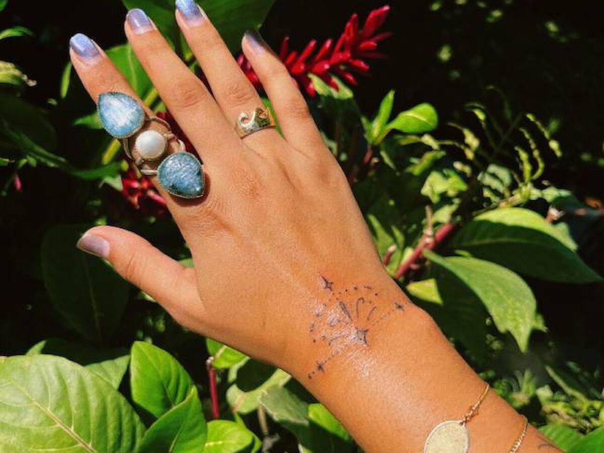 A hand adorned with multiple rings and bracelets, displaying a star and moon tattoo on the wrist, in front of lush green foliage.