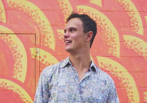 A person in a patterned shirt stands in front of a colorful, painted background featuring orange and yellow designs.