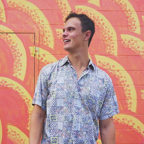 A person in a patterned shirt stands in front of a colorful, painted background featuring orange and yellow designs.