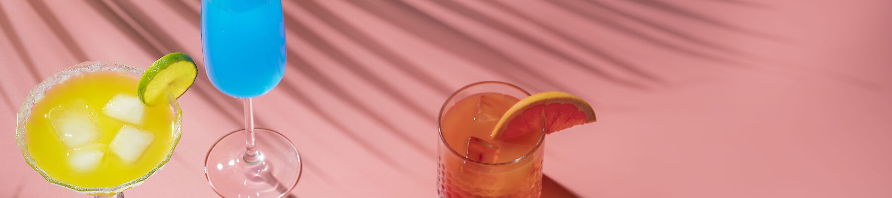 The image shows three colorful cocktails with citrus garnishes on a pink surface with palm leaf shadows cast over them.