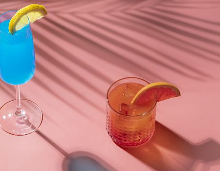 The image shows three colorful cocktails with citrus garnishes on a pink surface with palm leaf shadows cast over them.