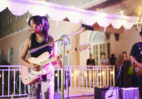 A woman plays an electric guitar onstage with lights overhead, while a man on the right also plays guitar. People in the background watch them.