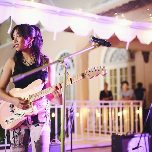 A woman plays an electric guitar onstage with lights overhead, while a man on the right also plays guitar. People in the background watch them.