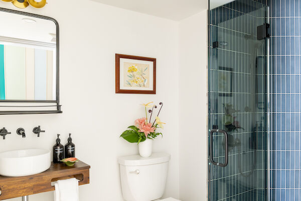 A bathroom with a mirror, sink, and toilet with flowers on top, and a framed picture on the wall next to a glass shower door ends the sentence.