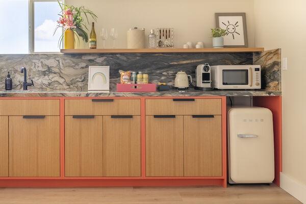 The image shows a modern kitchen with wooden cabinets, a marble backsplash, and various small appliances and decor, including a microwave and a mini-fridge.