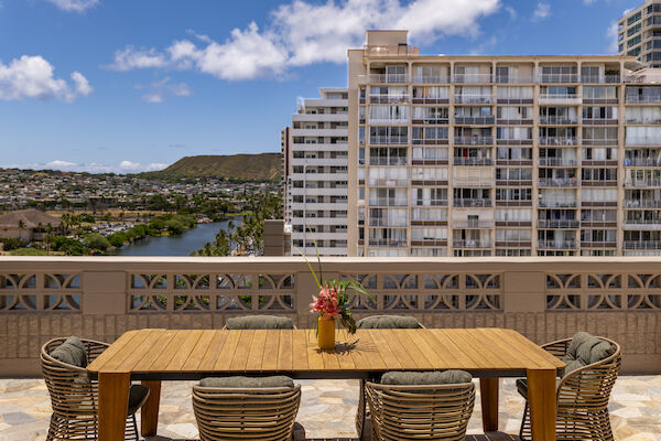 A rooftop terrace with a wooden table and six chairs overlooking a cityscape with buildings, a water body, and hills under a partly cloudy sky.