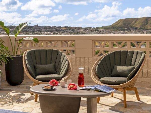 Two round chairs, a table with a jug, glasses, books, and red fruit, and a scenic view with mountains and blue sky are visible in the image.