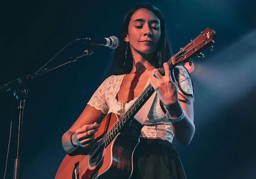 A woman is playing an acoustic guitar on stage under blue lighting, dressed casually in a white top and black skirt, in front of a microphone.