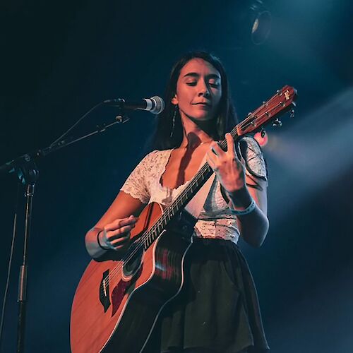 A woman is playing an acoustic guitar on stage under blue lighting, dressed casually in a white top and black skirt, in front of a microphone.