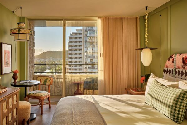 A cozy bedroom features a neatly made bed, a small dining table with chairs, and a large window with a balcony view of buildings and mountains.