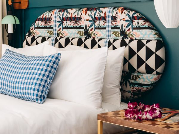 A bright bedroom: made bed with patterned pillows, a teal headboard, and a bedside lamp beside a small table with flowers.