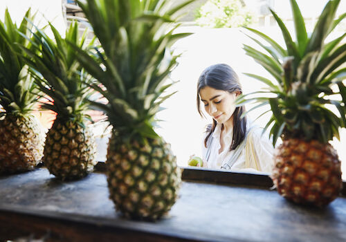 The image shows a woman standing behind a row of pineapples, focusing on something she is holding, with bright natural light in the background.