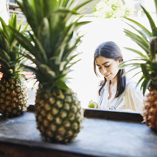 The image shows a woman standing behind a row of pineapples, focusing on something she is holding, with bright natural light in the background.