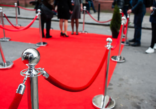 The image shows a red carpet event setup with stanchions and ropes, while people in formal attire stand in the background.