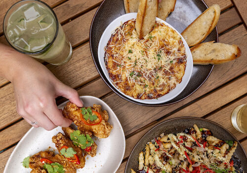 An assortment of dishes on a wooden table, including a pasta dish, bread, fried appetizers, and beverages. Hands are reaching for the food.