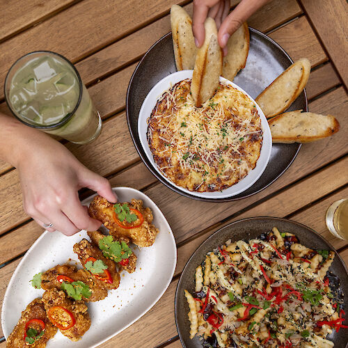 An assortment of dishes on a wooden table, including a pasta dish, bread, fried appetizers, and beverages. Hands are reaching for the food.