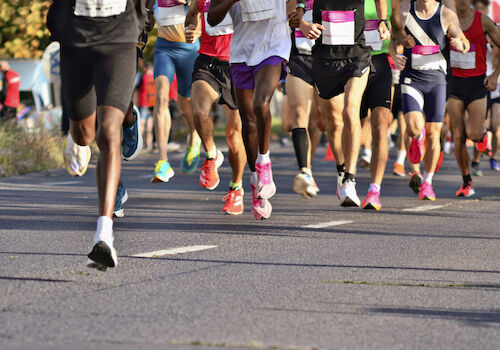 A group of runners participating in a race, seen from the legs down, with colorful running shoes and race bibs visible.