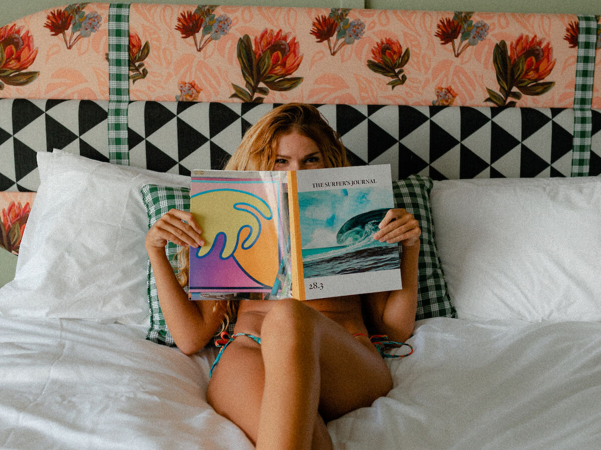 A person lying on a bed reading a colorful book, with a floral headboard behind them and white bed sheets.