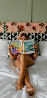 A person relaxing on a bed, reading a colorful book with a floral headboard in the background, dressed in casual, comfortable clothing.