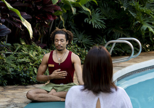 Two people are meditating by a pool surrounded by lush greenery.