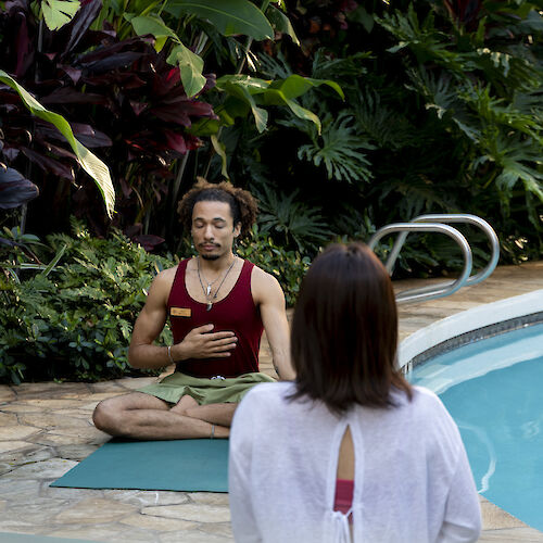 Two people are meditating by a pool surrounded by lush greenery.