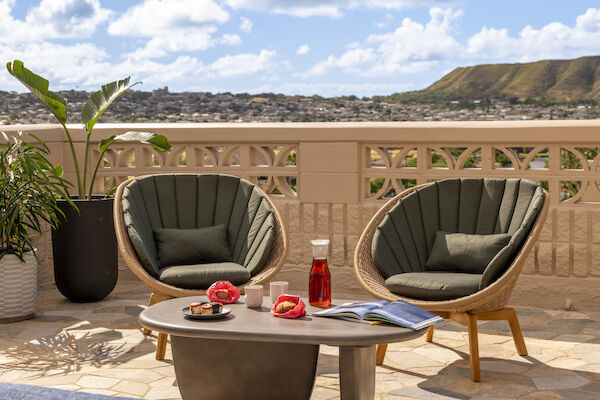 Two round chairs, a table with a jug, glasses, books, and red fruit, and a scenic view with mountains and blue sky are visible in the image.