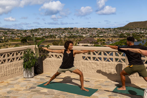 Two people are practicing yoga on a balcony on a sunny day with a scenic view in the background.