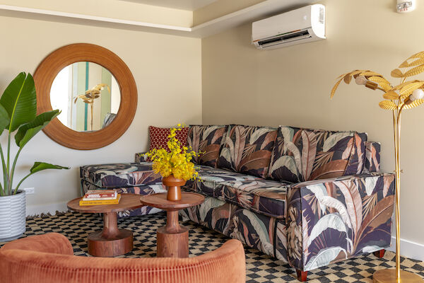 A cozy living room with a patterned sofa, round mirror, potted plants, coffee table, and cushions, adorned with warm tones and tropical decor.