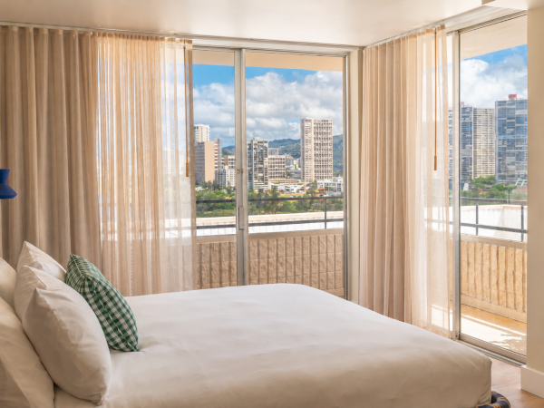 A bright hotel bedroom with a low bed, beige curtains, and large windows showing a city skyline under cloudy skies.