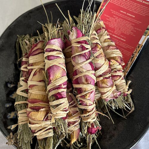 The image shows bundles of sage and rose petals wrapped with twine, sitting on a black plate with a red card nearby.