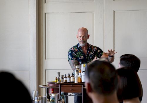 A bartender is preparing drinks in front of an audience, with bottles and ingredients on a cart in a room.