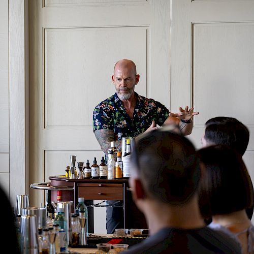 A bartender is preparing drinks in front of an audience, with bottles and ingredients on a cart in a room.