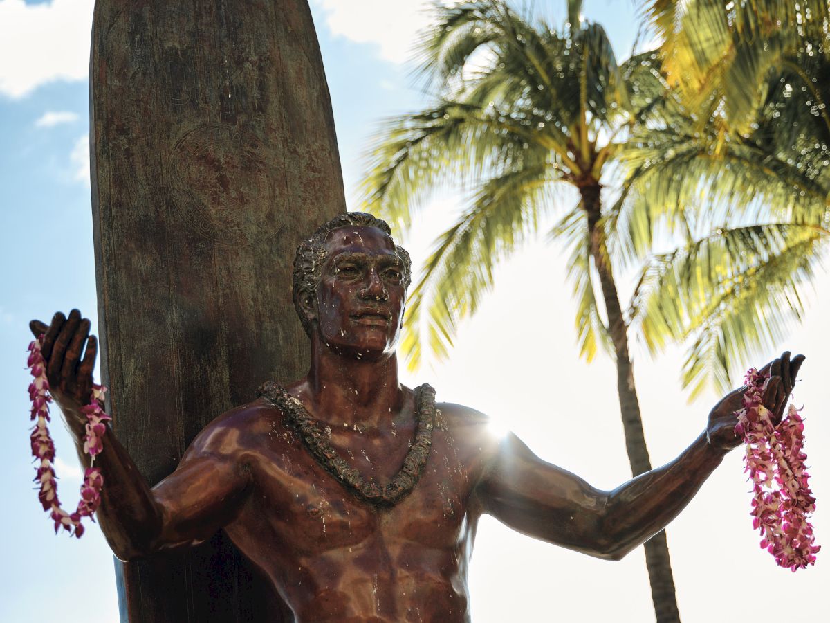 A statue of a surfer holding leis, with a surfboard behind, palm trees, and a sunny sky in the background.