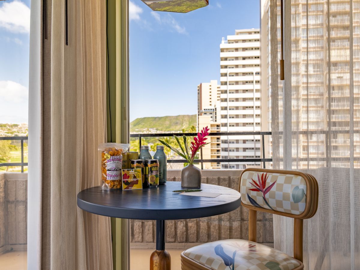 A small balcony with a round table and chair, snacks, drinks, and a vase with a flower, overlooking city buildings on a clear day.