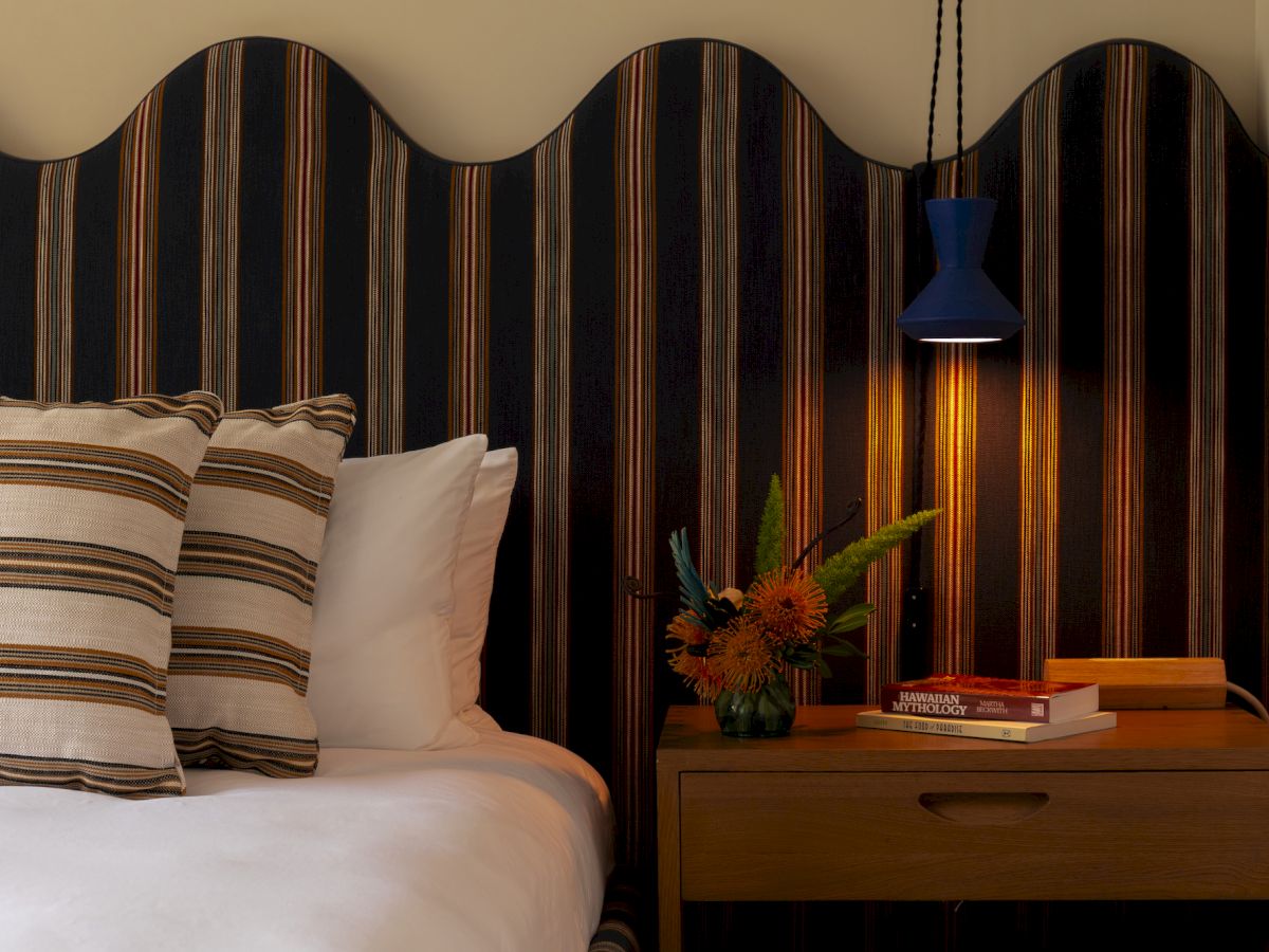 A cozy bedroom scene with a striped headboard, pillows, a wooden nightstand with a lamp, books, and a small floral arrangement.