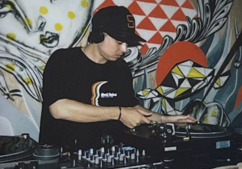 A DJ is mixing music on turntables in front of a colorful mural with abstract and geometric designs.