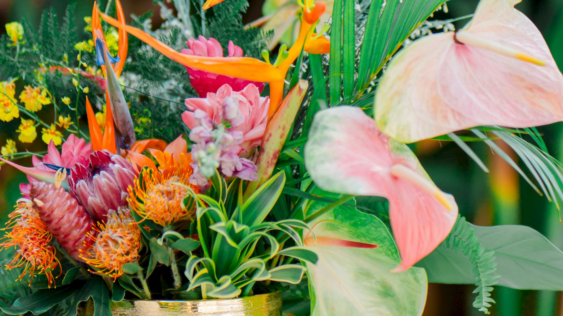 A colorful flower arrangement in a gold vase with various tropical flowers and greenery on a wooden stand outdoors.