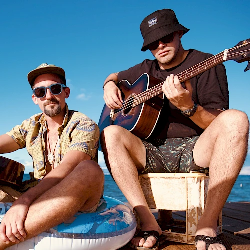 Two men sit outdoors by the water, playing guitars. They wear hats and sunglasses, embracing a casual, relaxed vibe under a clear sky.