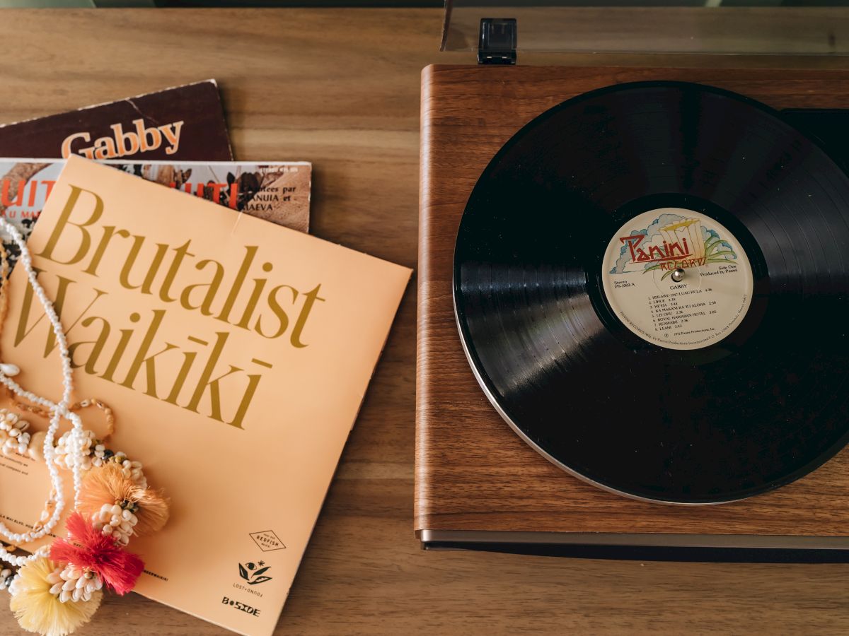 A wooden turntable with a spinning record is next to a few vinyl albums and a colorful lei necklace on a wooden surface.