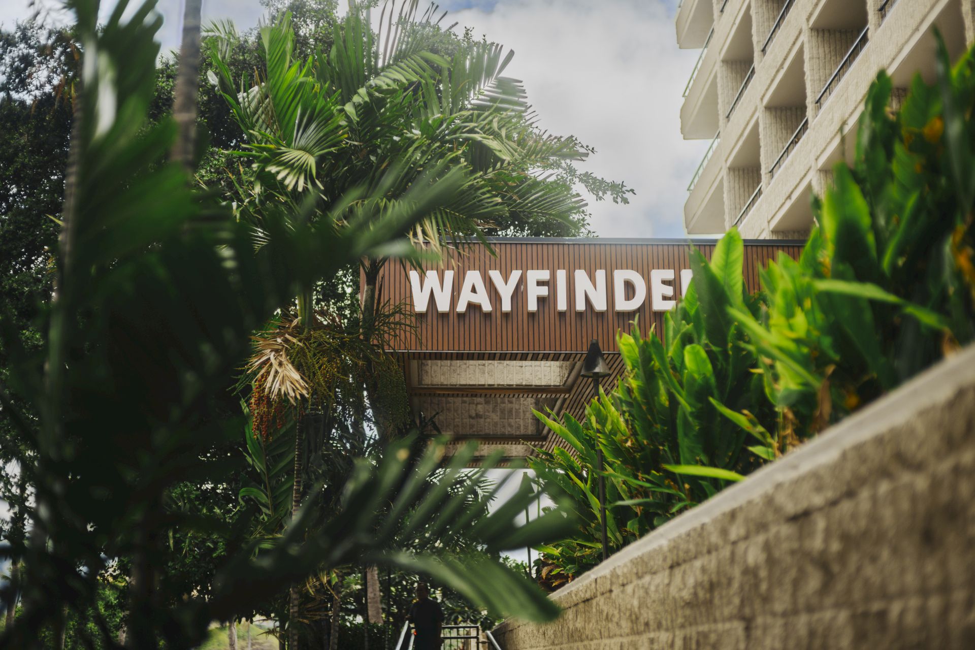 Un panneau indique &laquo; WAYFINDER &raquo; au milieu d'une v&eacute;g&eacute;tation tropicale luxuriante et d'un mur en b&eacute;ton &agrave; c&ocirc;t&eacute; d'un immeuble de plusieurs &eacute;tages, sugg&eacute;rant l'entr&eacute;e d'un complexe h&ocirc;telier ou d'un h&ocirc;tel.